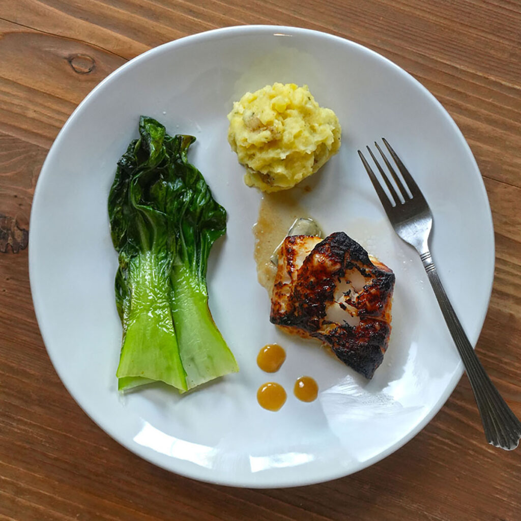 Pan‑seared black cod with a caramelized crust served alongside sautéed greens and a creamy mashed potato scoop on a white plate.