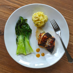 Pan‑seared black cod with a caramelized crust served alongside sautéed greens and a creamy mashed potato scoop on a white plate.