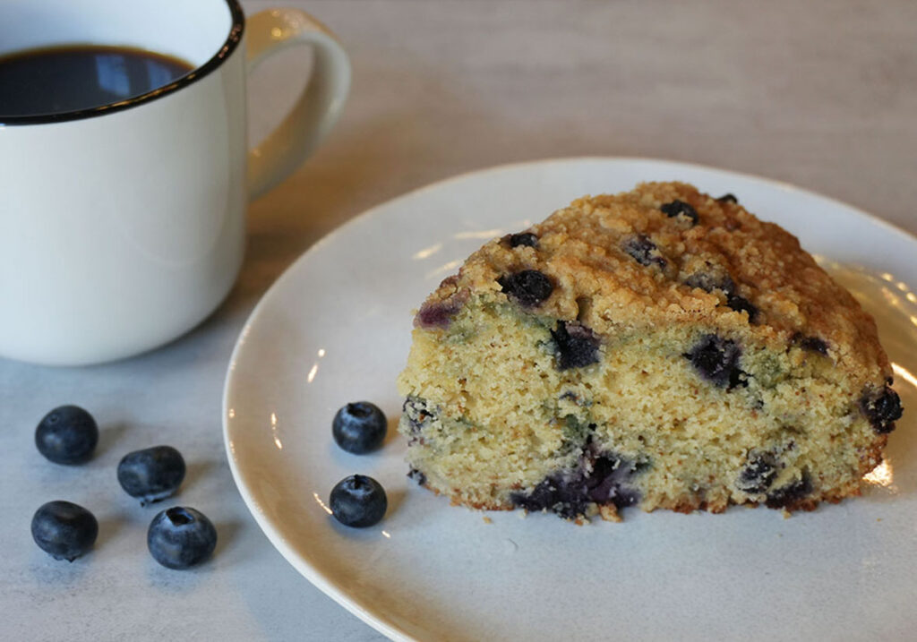 A slice of blueberry ricotta coffee cake sits on a white plate beside fresh blueberries, with a mug of coffee in the background on a light gray surface.