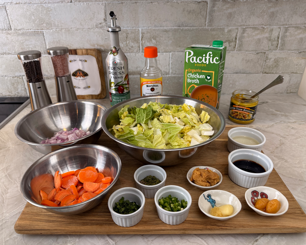 A neatly organized ingredient setup for ginger napa cabbage soup, featuring bowls of chopped carrots, onions, and napa cabbage surrounded by small dishes of garlic, ginger, green onions, chiles, miso, soy sauce, and seasonings on a wooden board.