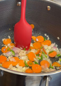 Vegetables sautéing in a pan—carrots, onions, garlic, and chiles being stirred with a red spatula during the first step of cooking.