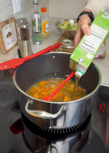 Broth being poured into a pot of sautéed vegetables while stirring with a red spatula during the second step of cooking.