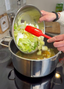 Chopped napa cabbage and vegetables being poured from a metal bowl into a pot of simmering broth while stirring with a red spatula.