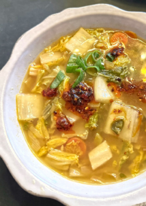 A bowl of ginger napa cabbage soup filled with napa cabbage, carrots, green onions, and chili paste in a light golden broth.