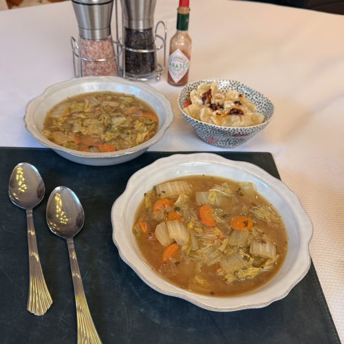 Two bowls of ginger napa cabbage soup on a woven placemat with two spoons, featuring dumplings, Tabasco sauce, and a salt and pepper grinder.