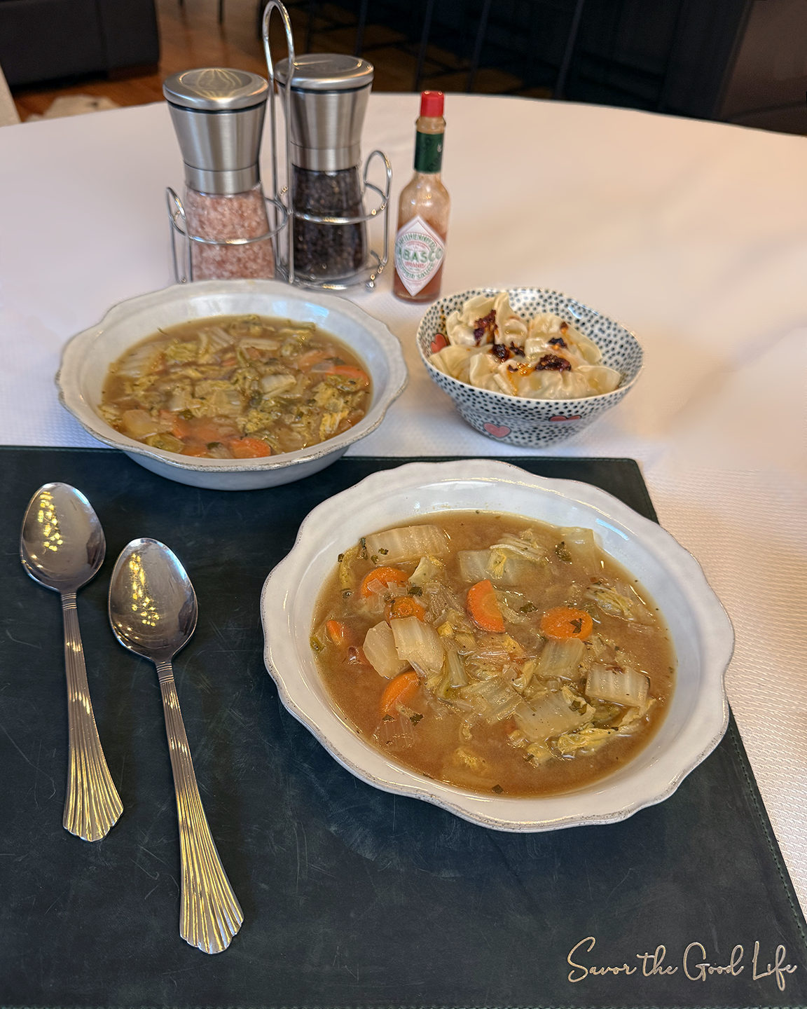 Two bowls of ginger napa cabbage soup on a woven placemat with two spoons, featuring dumplings, Tabasco sauce, and a salt and pepper grinder.