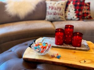A glass bowl filled with colorful candies on a wooden coffee table beside red candle holders, with a sofa and a “Merry Christmas” pillow in the background.