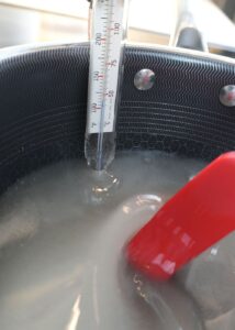 A pot of heating sugar syrup with a candy thermometer inserted and a red spatula resting inside, showing the temperature‑monitoring stage of hard‑candy making.