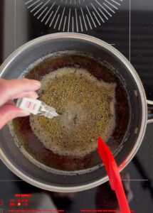 A hand holding a red silicone spatula above a bubbling pot of hot sugar syrup with a red spatula inside, showing the moment just before adding LorAnn flavoring in Step 4 of hard‑candy making.