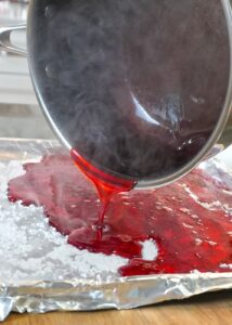 A pot of hot red candy syrup being poured onto a foil‑lined sheet pan dusted with confectioners’ sugar during the hard‑candy making process.