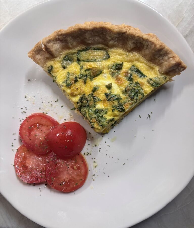 Slice of baked quiche on a white plate with fresh tomato slices.