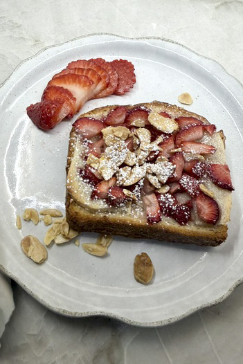 A full strawberry almond croissant toast on a cream-colored plate, topped with fresh sliced strawberries, sliced almonds, and a dusting of powdered sugar, with additional strawberries and almonds scattered around the plate.