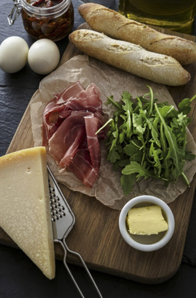 Prosciutto, arugula, parmesan wedge, and sliced baguette arranged on a wooden board for sandwich prep.
