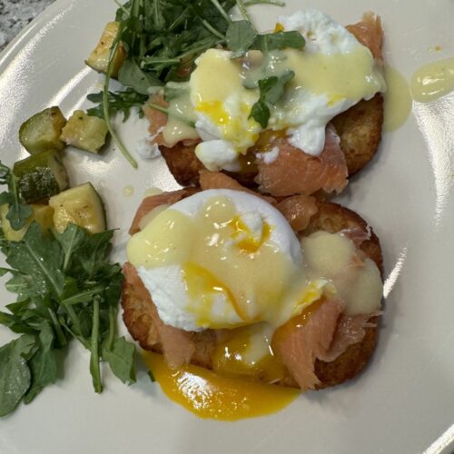 Eggs Benedict with lox served on crispy hashbrowns with hollandaise and a fresh side salad on a plate.