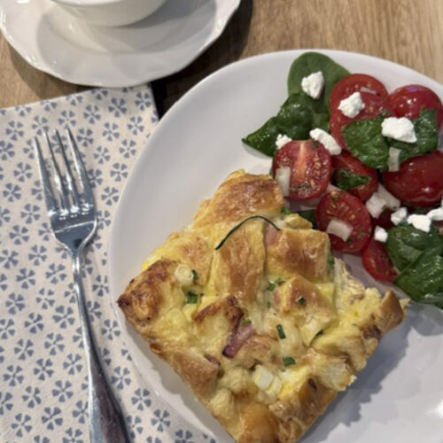 Hero plate with ham and Swiss croissant bake served alongside a fresh side salad and sautéed spinach.