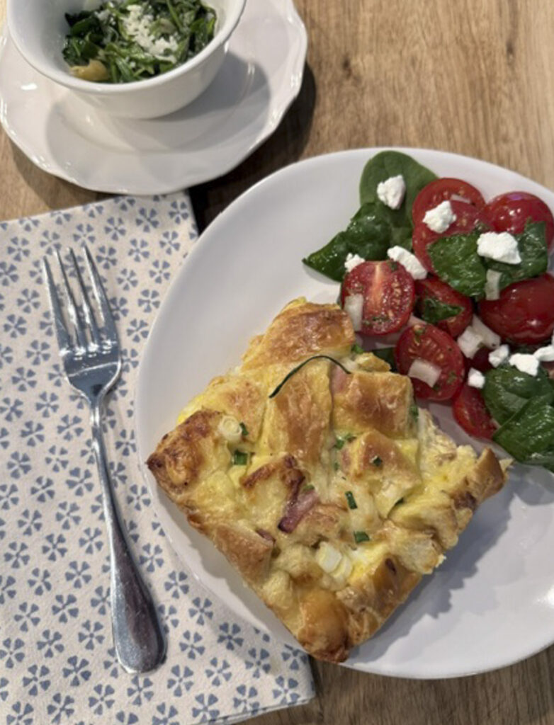 Hero plate with ham and Swiss croissant bake served alongside a fresh side salad and sautéed spinach.