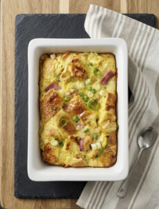 Golden ham and Swiss croissant bake in a dish after baking, showing crisp layered croissants during the rest period.