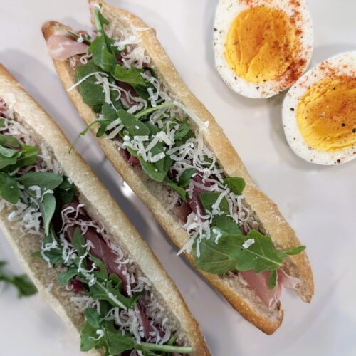 Mini baguette sandwiches with prosciutto, arugula, and shaved Parmesan on a plate, served alongside a hard‑boiled egg cut in half.