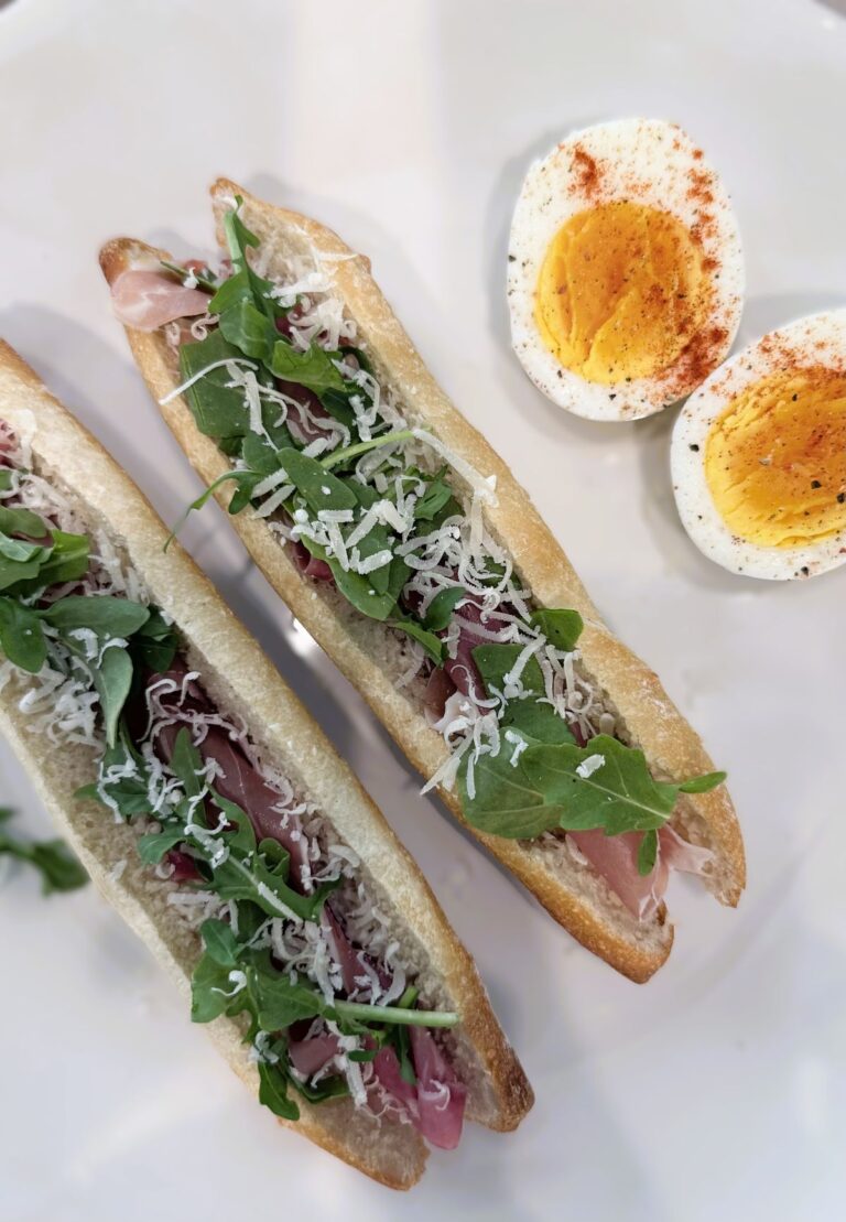 Mini baguette sandwiches with prosciutto, arugula, and shaved Parmesan on a plate, served alongside a hard‑boiled egg cut in half.
