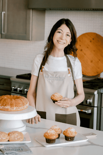 Samantha wearing a Full Bloom Bites apron in her kitchen, holding a fresh muffin beside a display of homemade pastries.